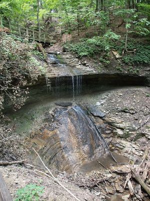 [A wide but small volume water stream spills off a ledge about 6 feet above the lower rock. It then runs down a rock face approximately 15 feet long before becoming a stream again.]